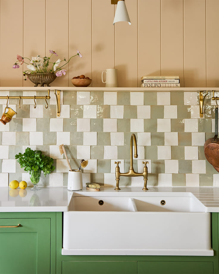 Charming kitchen with leafy green lower cabinets, checkered square zellige tiles in cream and sage, brass tapware, and a farmhouse sink