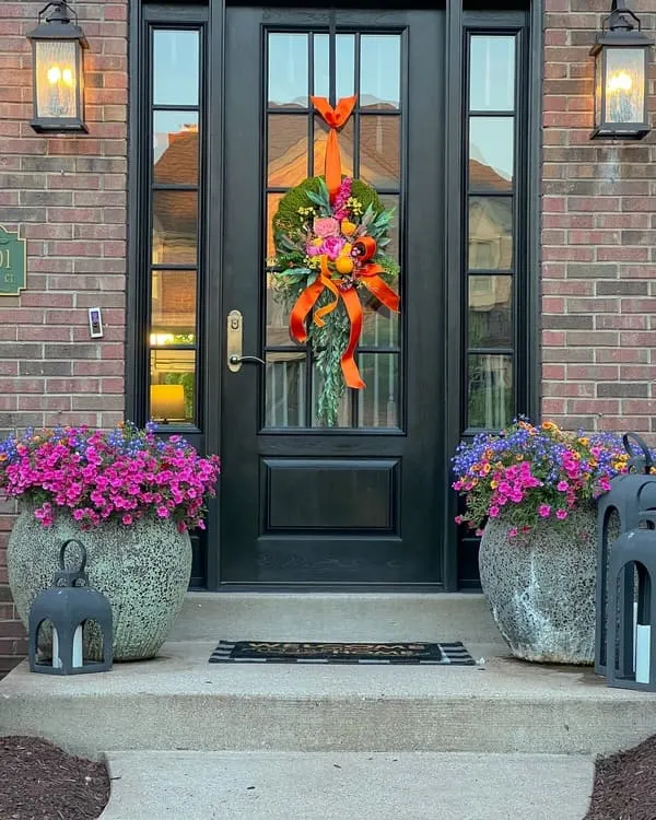 Brick facade dotted with a black door, bright floral wreath and colorful planters.