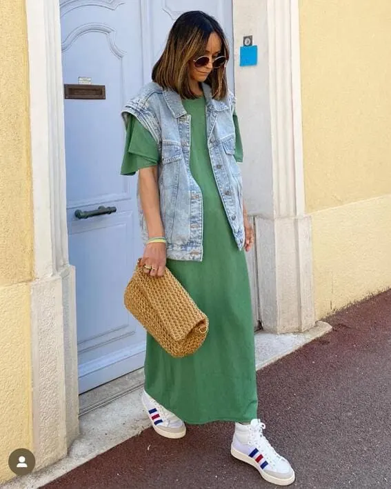 A woman wearing an denim vest, worn over a maxi dress in green with sneakers.