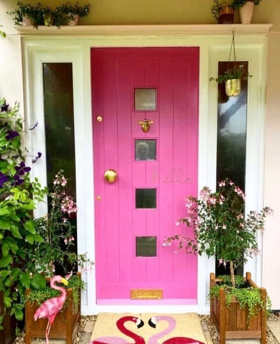 Bright pink front door with golden accents, framed by white trim and surrounded by potted plants and flamingo decor.