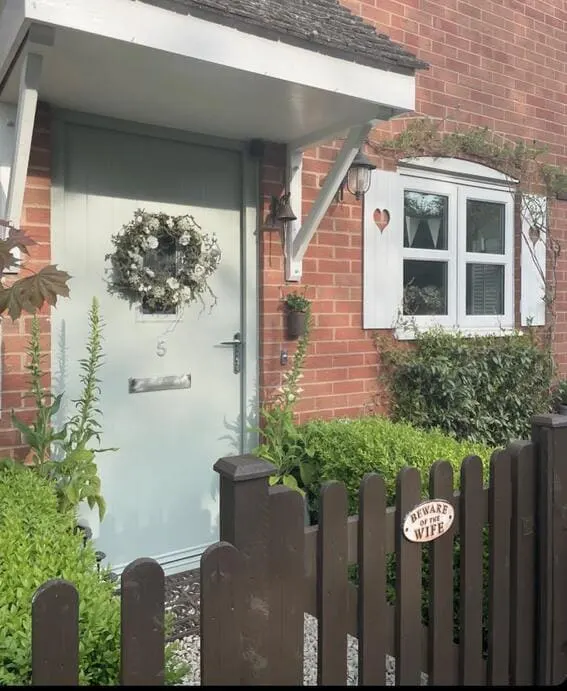 Soft green front door with a floral wreath, framed by a small garden and wooden gate.
