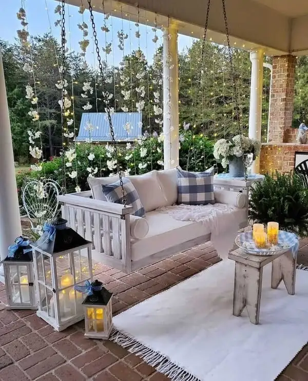 A dreamy porch featuring a white hanging daybed, blue gingham pillows, overflowing floral garlands, flickering lanterns, and a white fringed rug.