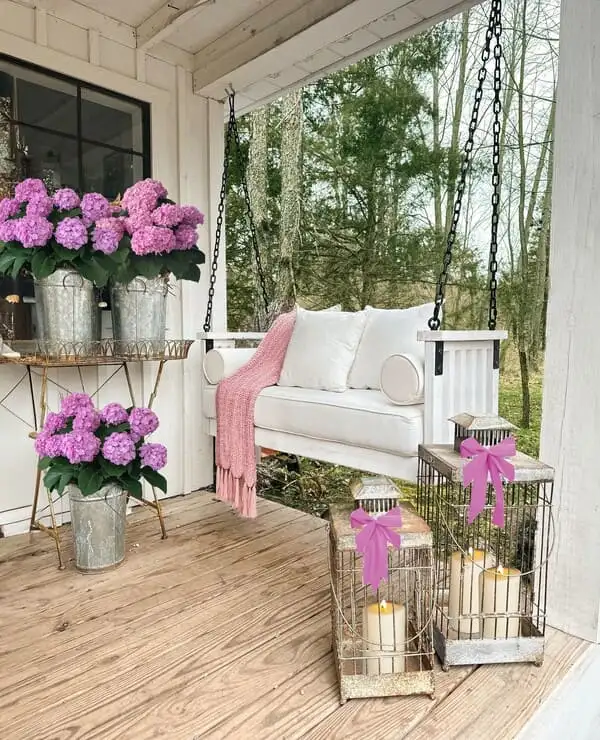 A farmhouse-style porch with a white swing, galvanized planters of pink hydrangeas, wire lanterns with bows and a blush throw blanket.