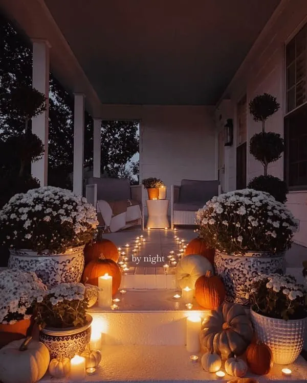 A moody fall porch illuminated with candlelight, pumpkins, mums in chinoiserie planters and soft seating at the far end.