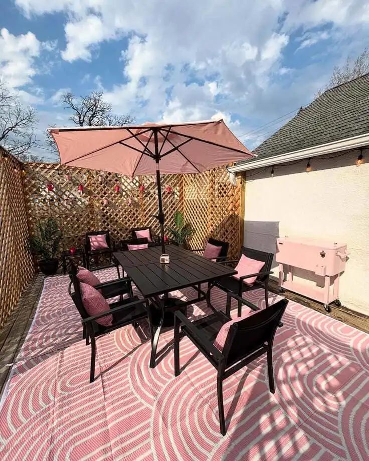 A modern outdoor patio featuring a pink and white patterned rug, black dining set, pink pillows, pink umbrella, and a pink cooler.