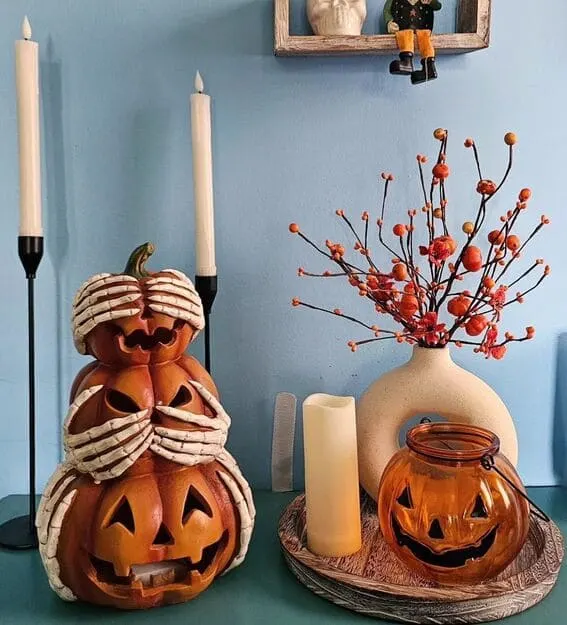 A quirky and charming vignette featuring stacked jack-o'-lanterns with skeleton hands, paired with a classic pumpkin lantern and vibrant orange berry branches.