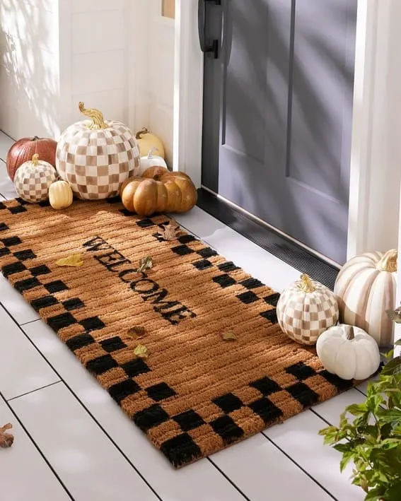 A cozy front porch setup featuring checkered and neutral-toned pumpkins beside a welcoming doormat.